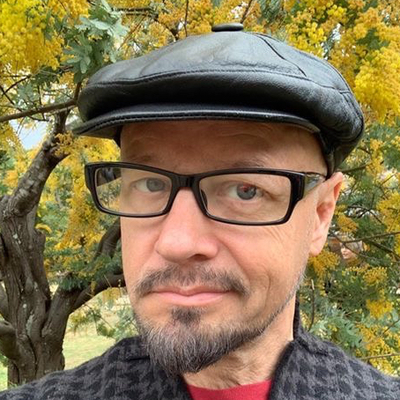 Portrait of Phil Stilwell wearing glasses and a black cap in front of yellow blossoms