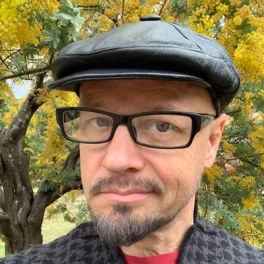 Portrait of Phil Stilwell wearing glasses and a black cap in front of yellow blossoms